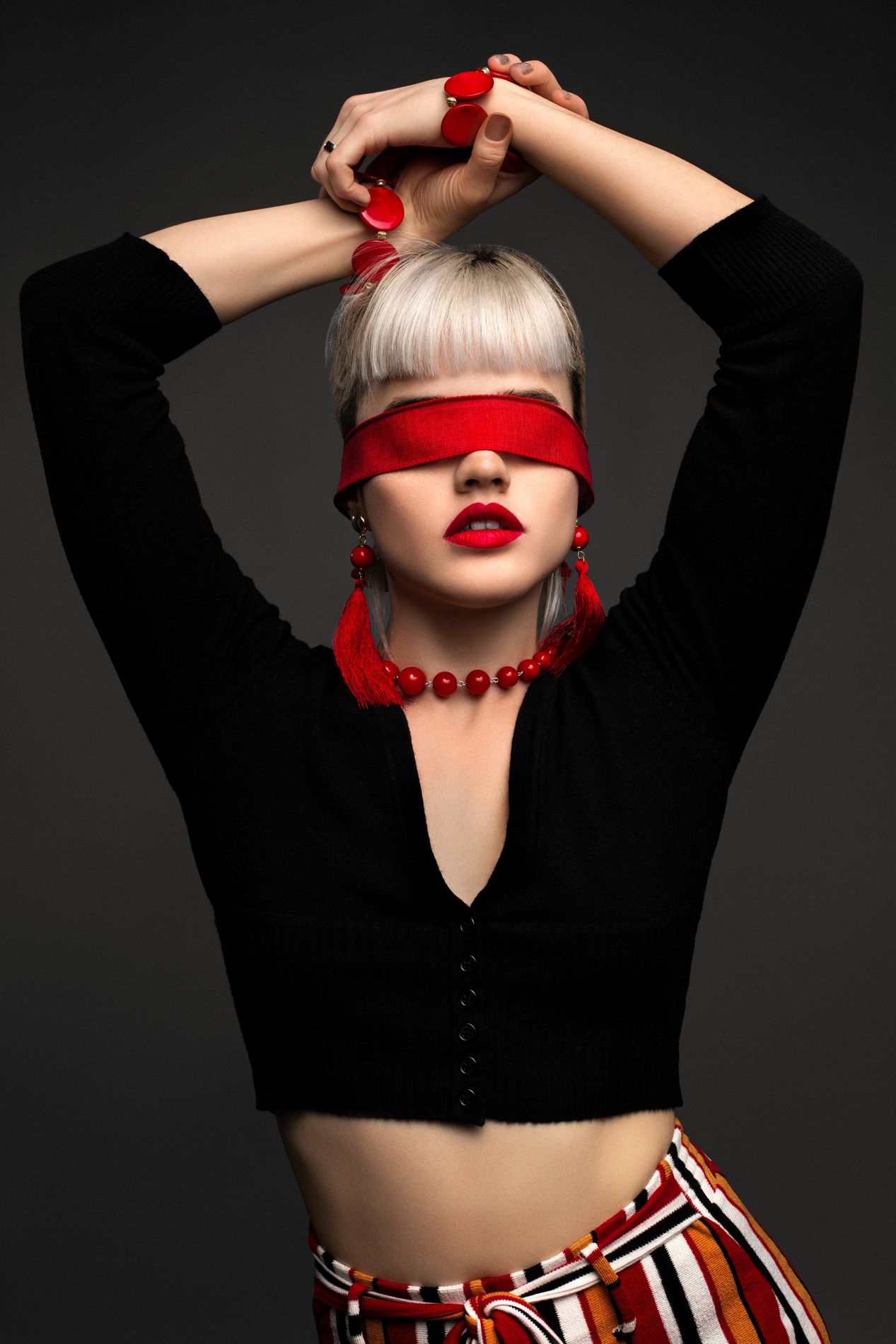Studio fashion portrait of Violet in a black top, striped trousers, red jewelry, and a red eye covering.